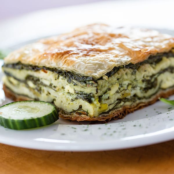 Close-up of a slice of spanakopita with flaky crust, spinach, and cheese, garnished with cucumber.