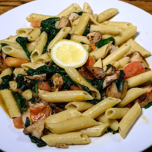 Penne pasta with chicken, spinach, tomatoes, and a lemon slice on a white plate.