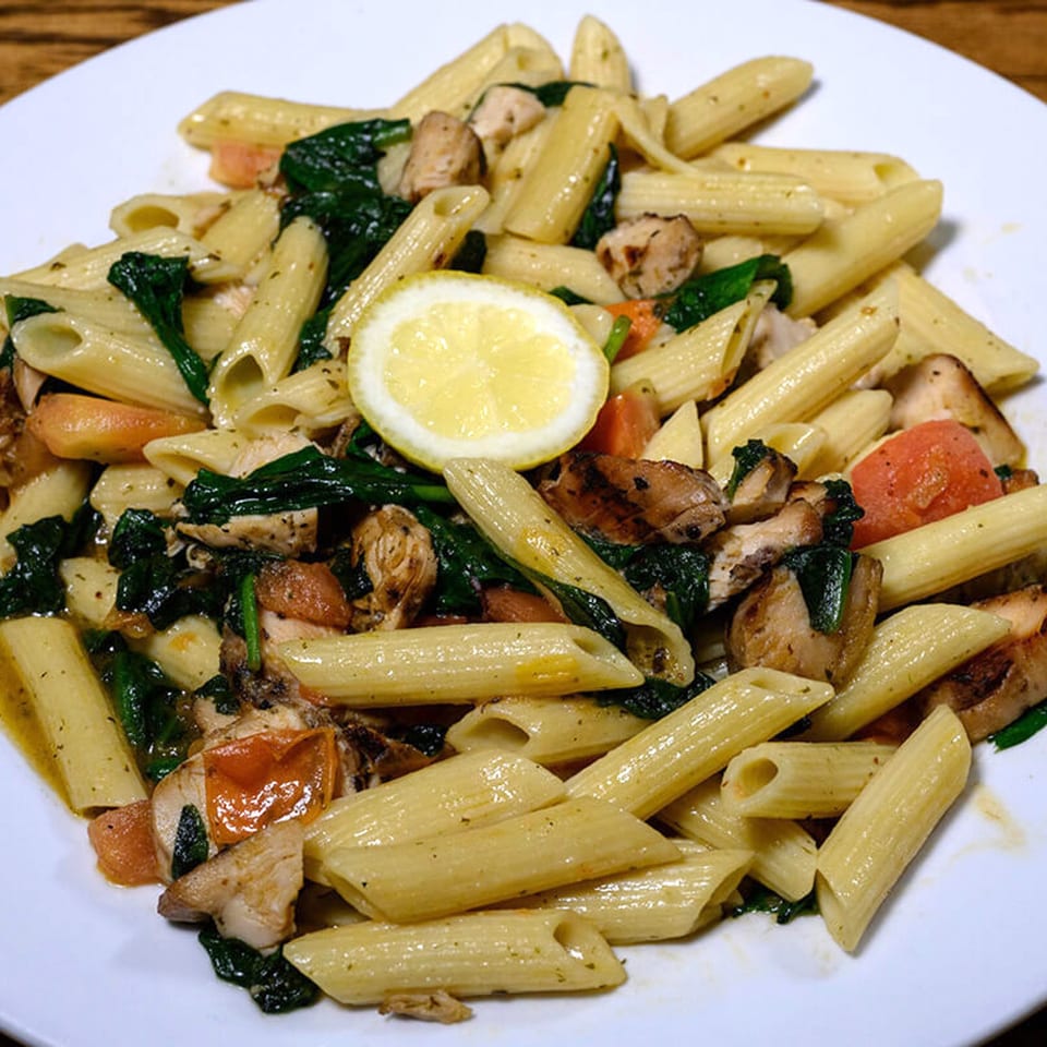 Penne pasta with chicken, spinach, tomatoes, and a lemon slice on a white plate.