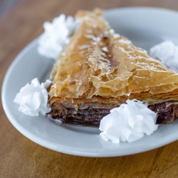 A triangular slice of baklava with flaky pastry layers and whipped cream on a white plate.