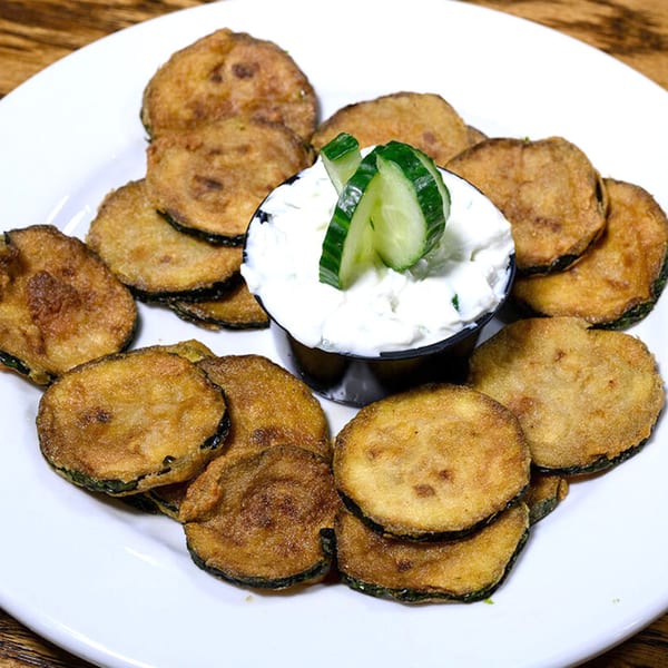 Crispy fried zucchini slices with a side of tzatziki sauce, garnished with cucumber, on a white plate.
